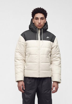 Joven con chaqueta acolchada Nike en negro y blanco con capucha, de pie frente a un fondo claro y liso.