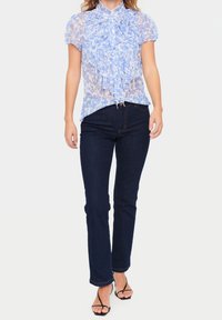 Blouse transparente bleu clair avec un col à volants et un devant froncé, associée à un jean évasé bleu foncé. La tenue est complétée par des sandales noires à lanières.