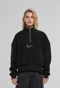 Pullover in pile nero con colletto alto, chiusura a mezza zip e logo bianco ricamato sul davanti; presenta polsini e orlo a costine.
