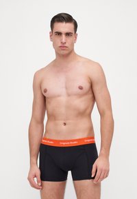 Zwarte boxershorts met een oranje tailleband met de tekst "Originals Studio". De stof lijkt glad met een aansluitend ontwerp.