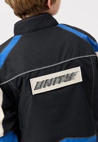 Svart jacka med blå och vita accenter, försedd med en stor vit patch med "UNITY" i svart fetstil på baksidan.