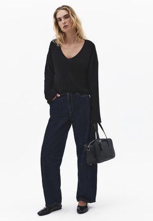 Kvinde iført sort V-hals sweater, mørke vide jeans, sorte flade sko og bærende en sort håndtaske med korte håndtag.