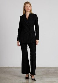 Zwart gestructureerd pak met een aansluitende blazer met een dubbelgebouwde ontwerp en bijpassende flared broek. Zwarte hoge hakken.