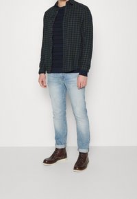 Codzienny strój składający się z zielono-granatowej koszuli w kratę, ciemnego swetra z dzianiny, jasnoniebieskich dżinsów oraz brązowych skórzanych butów na białej podeszwie.