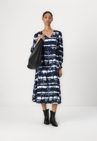 Blå- og hvit tie-dye-kjole med lange ermer, V-hals og en flott silhuett, sammen med svarte ankelstøvler og en stor svart veske.