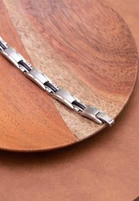 Zilveren metalen armband met gestructureerde schakels en zwarte accenten, rustend op een ronde houten schaal met een natuurlijke afwerking.