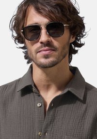 Grau strukturiertes Hemd mit sichtbaren Nähten, getragen unter einer schwarzen Sonnenbrille mit runder Fassung und dunklen Gläsern.