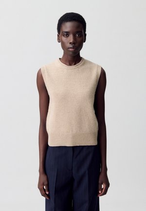 Tops en tricot sans manches beige avec ourlet côtelé et encolure ronde ; texture douce et design minimaliste. Associé à un pantalon sombre et bien ajusté.