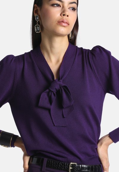 Femme portant un haut violet à manches longues avec un nœud noué au cou, de grandes boucles d’oreilles noires et une ceinture noire à texture croco.