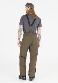 Mand med langt hår og tatoveringer iført brune snebukser med sorte seler, grå t-shirt, skistøvler og grønne skibriller på hovedet, der står med ryggen til.