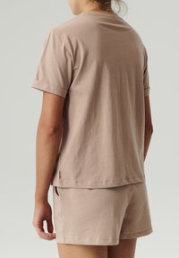 Kobieta w beżowej koszulce z krótkim rękawem i pasujących szortach, pokazana od tyłu na jednolitym jasnym tle.