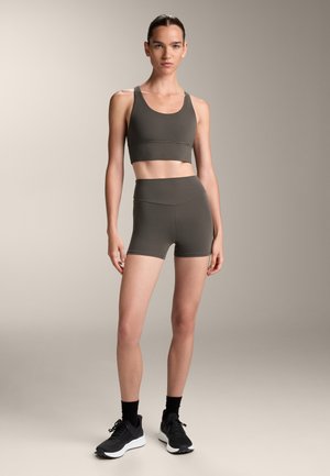 Olijf groene sportbeha en high-waisted shorts van rekbaar materiaal, met een naadloos ontwerp. Model draagt zwarte sneakers met sokken.