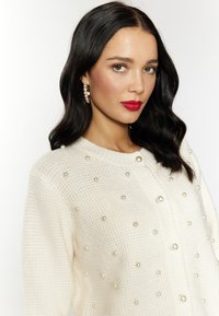Witte gebreide cardigan met textuurstof en grote decoratieve parels. Sluit aan de voorkant met een ronde halslijn en subtiele knoopdetails.
