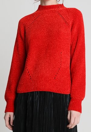 Kobieta ubrana w jasnoczerwony dzianinowy sweter z subtelnymi wzorami w połączeniu z czarną plisowaną spódnicą, stojąca na jednolitym tle.