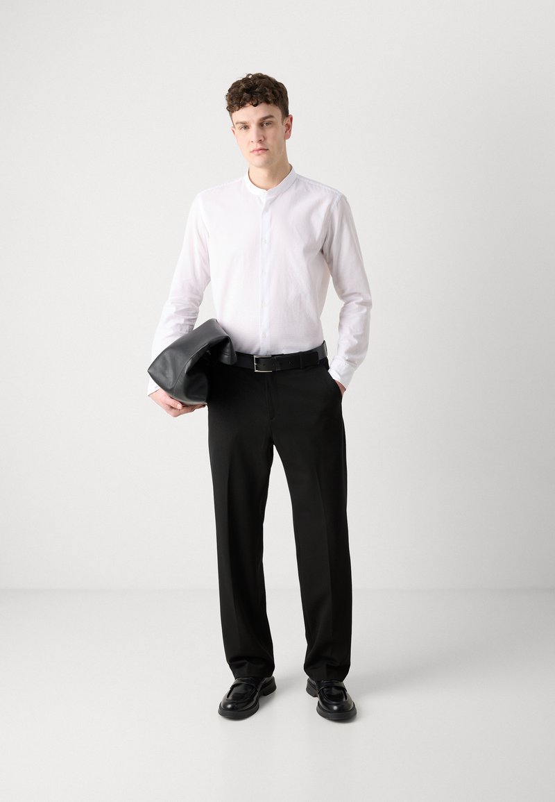 Biała koszula zapinana na guziki z kołnierzykiem mandaryńskim, w połączeniu z czarnymi spodniami. Model trzyma czarną skórzaną torbę i nosi czarne buty.