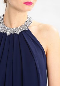 Navy halterklänning med veck, prydd med en glittrande kristallhalsringning. Slät tygliknande textur, elegant design utan ytterligare mönster.