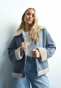 Jeansjacka med krämfärgad sherpa-krage och manschetter, med dragkedja framtill, avslappnad passform och blå färg. Modell som bär ljusa jeans och en vit topp.