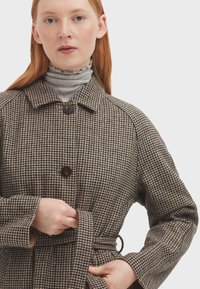 Abrigo de espiga marrón con grandes botones, un cuello y una cintura ceñida con cinturón. La tela texturizada resalta la artesanía y el diseño a medida.