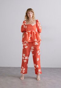 Rød blomstret peplum-topp med puffede ermer, parret med matchende vide bukser, begge med hvite blomstermønstre på et glatt stoff.
