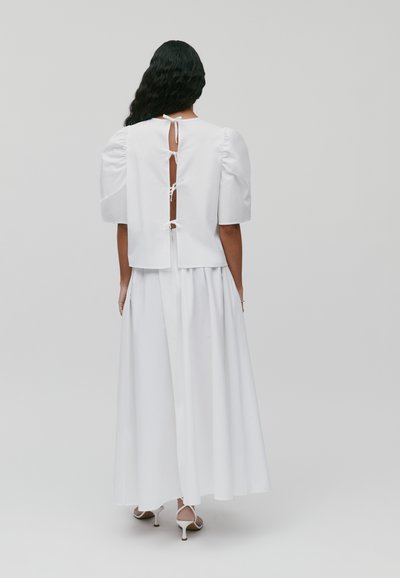 Weißes Kleid mit Puffärmeln und offenem Rücken, verziert mit Bandbindungen. Der Stoff ist glatt und bietet eine lockere Passform, die bis zum Knöchel fließt.