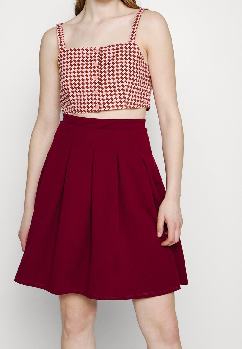 Femme portant un haut court sans manches avec un motif marron et rose et une jupe plissée bordeaux taille haute sur un fond uni.