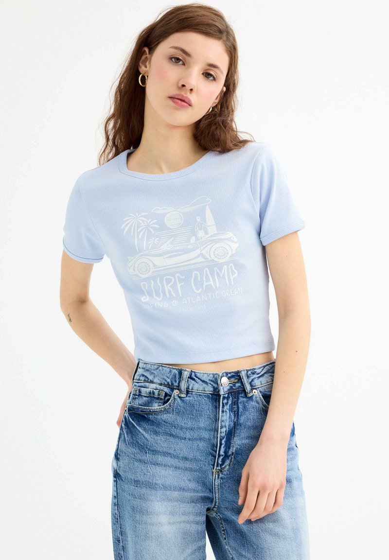 Camiseta corta azul claro con mangas cortas y textura acanalada, que presenta un gráfico de campamento de surf blanco y un diseño de palmera en la parte delantera.