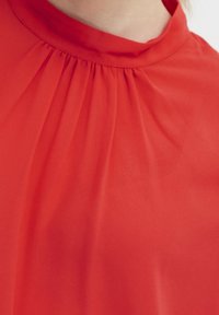 Rote Bluse mit einem hohen, gerafften Ausschnitt und weichem, fließendem Stoff mit glatter Textur. Keine sichtbaren Muster oder Akzente.