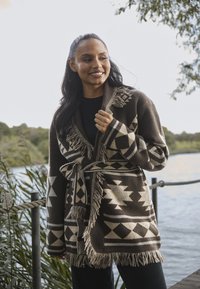 Bruine gebloemde cardigan met franjes en een riem. Kenmerkt zich door geometrische ontwerpen in roomwit op een donkere achtergrond. Zachte textuur.