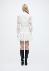 Γυναίκα στέκεται πλάτη φορώντας λευκό δαντελένιο φόρεμα με φουσκωτά μανίκια και μαύρες ψηλές μπότες μέχρι το γόνατο, σε απλό λευκό φόντο.