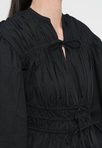 Svart bluse med teksturert, plissert stoff. Har kragefri halslinje, frontbånddetalj og samlet elastisk midje. Lange ermer.