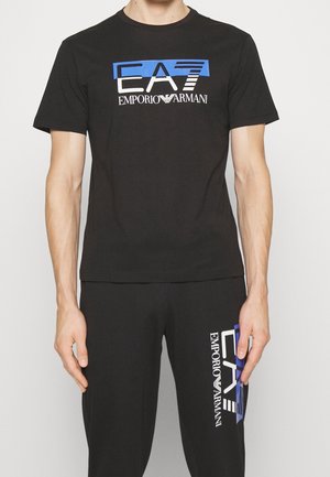 Mężczyzna ubrany w czarną koszulkę i spodnie z niebiesko-białymi logo „EA7 Emporio Armani” na piersi i udzie.