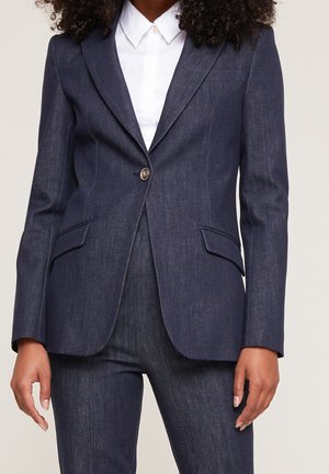 Woman wearing dark denim fitted blazer with single button and matching pants, paired with white collared shirt.