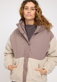 Brown and beige hooded jacket with soft fleece texture. Features snap buttons, side pockets, and a two-tone design. Brand logo visible.