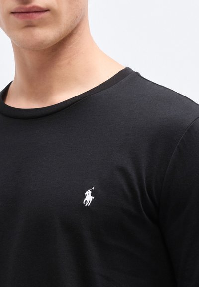 Svart T-skjorte med rund hals, laget av bomull. Har en liten hvit polo-spillerlogo på venstre side av brystet. Jevn tekstur.