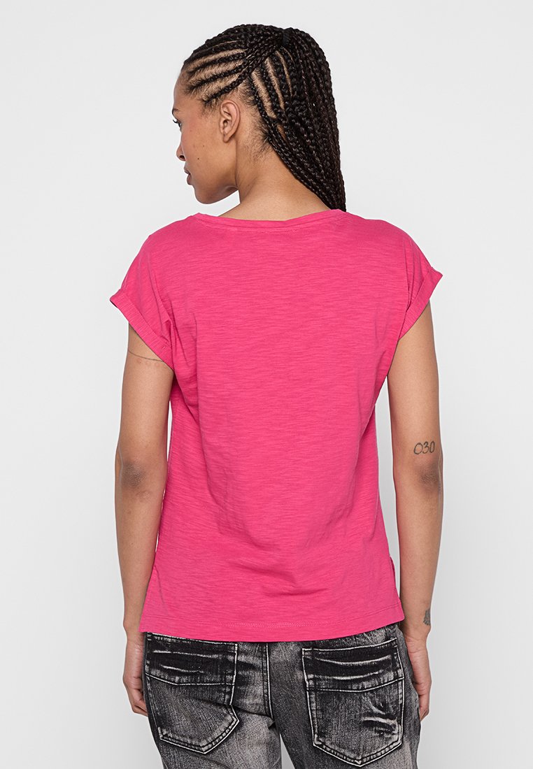Frau mit geflochtenem Haar, die ein leuchtend pinkes kurzärmliges Shirt und ausgewaschene schwarze Jeans trägt, von hinten vor einem weißen Hintergrund gezeigt.