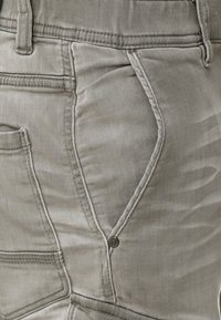 Lichtgrijze denim broek heeft een gestructureerd oppervlak, flapzakontwerp en accentuering met topstitching, met een klein metalen knoopdetail.