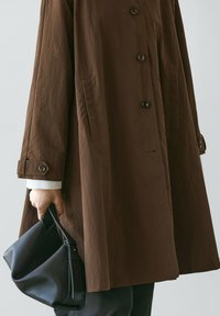 Person in brown buttoned coat holding a black leather handbag by its handle at their side against a plain background.