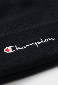 Berretto di maglia nera con logo "Champion" ricamato bianco, arricchito da un logo distintivo in rosso e bianco. Dettagli del tessuto testurizzato visibili.