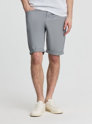 Mężczyzna ubrany w jasnoszorty z podwiniętymi brzegami, białe sportowe buty i białą koszulę, stojący z jedną ręką w kieszeni na jednolitym jasnym tle.