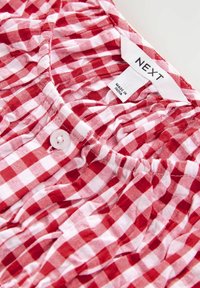 Red and white checked fabric, lightweight textile with pleats, featuring a white button and a label showing "NEXT, MADE IN INDIA."