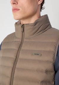 Bruine puffer gilet met hoge kraag en ritssluiting. Heeft horizontale stiknaden en een Calvin Klein logo label op de voorkant. Getextureerd stof.