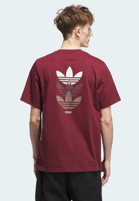 Camiseta de algodón color burdeos con un gran gráfico en capas de logotipos de trébol de Adidas en blanco en la parte posterior. Diseño de manga corta y cuello redondo.