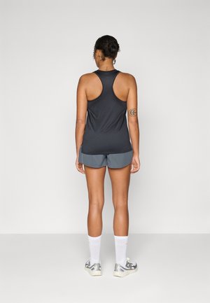 Frau, die mit dem Rücken zur Kamera steht, trägt ein schwarzes Racerback-Top, graue Shorts, weiße Kniestrümpfe und graue Sneakers vor einem schlichten Hintergrund.