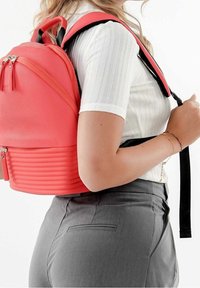 Zaino color corallo con design testurizzato, chiusura con cerniera e costine orizzontali nella parte inferiore. Spallacci neri e dettagli hardware visibili.