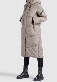 Beige Steppjacke mit Kapuze, diagonal genäht, mit Reißverschlusstaschen und Druckknöpfen am Saum. Getragen über schwarzen Hosen und Stiefeln.