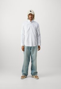 Camicia bianca a maniche lunghe, jeans a gamba larga azzurri chiari, berretto beige con logo LA e scarpe beige-marroni con suole bianche. Abbigliamento semplice e informale.