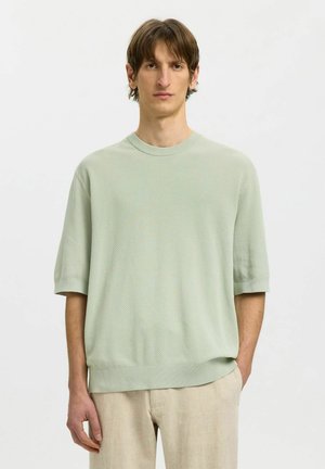 Homme debout portant un pull texturé à manches courtes vert pâle et un pantalon beige sur un fond clair uni.