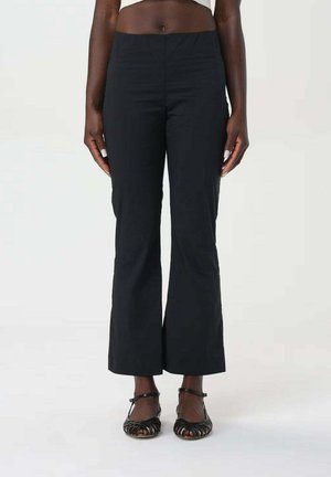 Zwart slim-fit broek met lichte uitlopende pijpen gedragen door een staande persoon, gecombineerd met zwarte platte sandalen met bandjes.