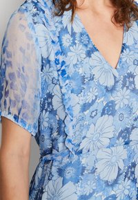 Robe florale bleu clair avec un col froncé et des manches courtes et translucides. Le tissu présente divers motifs de fleurs dans des nuances de bleu.