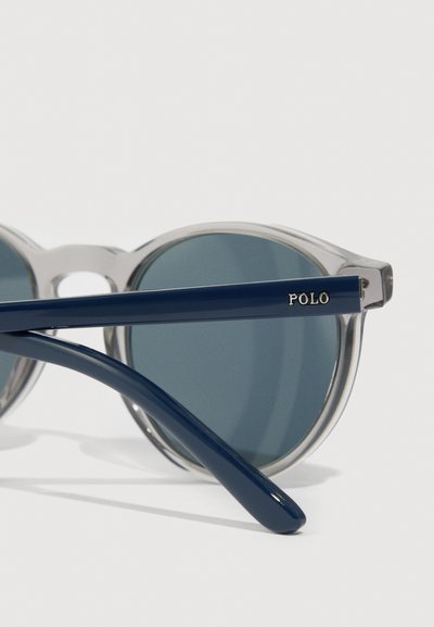 Ochelari de soare rotunzi cu ramă din plastic transparent și brațe de culoare albastră închisă. Lentilele sunt nuanțate în gri, cu "POLO" gravat pe accentele brațelor.